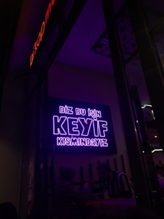 A neon sign in bright purple lettering with the text 'Biz bu işin keyif kısmındayız' is mounted on the wall. Below the sign, two people are seated together in a dimly-lit setting, suggesting a cozy atmosphere.