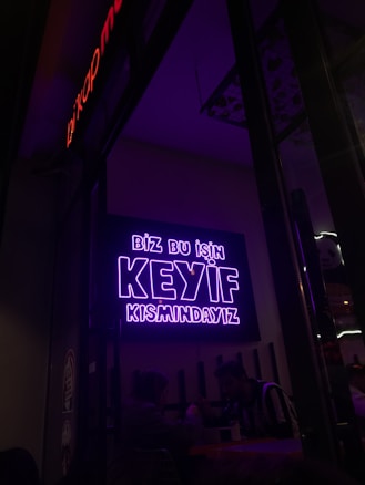 A neon sign in bright purple lettering with the text 'Biz bu işin keyif kısmındayız' is mounted on the wall. Below the sign, two people are seated together in a dimly-lit setting, suggesting a cozy atmosphere.