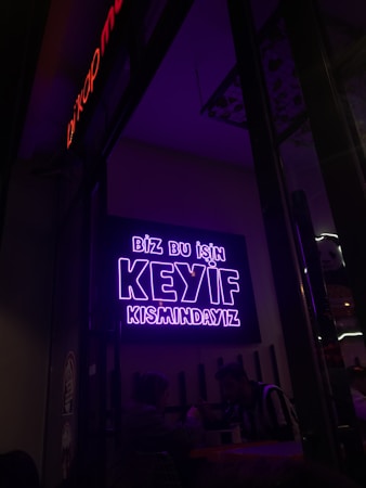 A neon sign in bright purple lettering with the text 'Biz bu işin keyif kısmındayız' is mounted on the wall. Below the sign, two people are seated together in a dimly-lit setting, suggesting a cozy atmosphere.