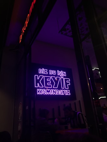 A neon sign in bright purple lettering with the text 'Biz bu işin keyif kısmındayız' is mounted on the wall. Below the sign, two people are seated together in a dimly-lit setting, suggesting a cozy atmosphere.