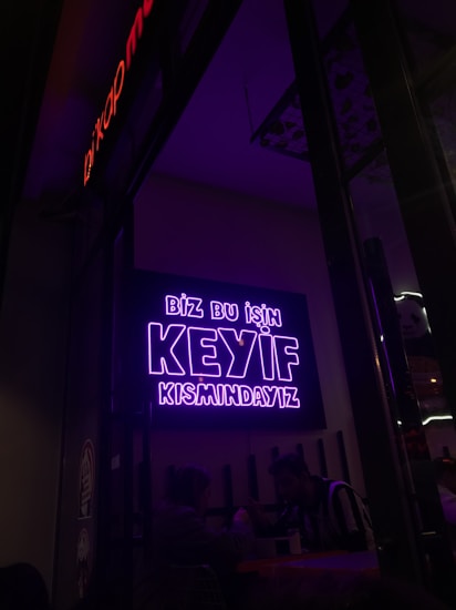 A neon sign in bright purple lettering with the text 'Biz bu işin keyif kısmındayız' is mounted on the wall. Below the sign, two people are seated together in a dimly-lit setting, suggesting a cozy atmosphere.