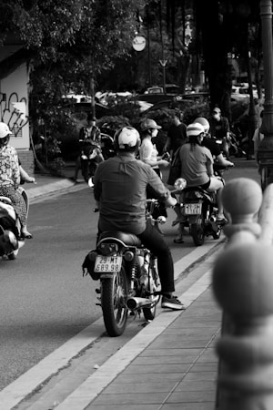 A black and white scene of people riding motorcycles on a street, possibly in an urban environment. Some riders are wearing helmets and casual clothing. Trees and parked cars line the background, while a pavement runs along one side of the road.