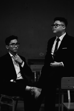 Two people are sitting together in a dimly lit room. They are dressed in formal attire, both wearing dark suits and glasses. One person is sitting with a thoughtful expression, adjusting their tie, while the other sits relaxed, resting one leg over the other.