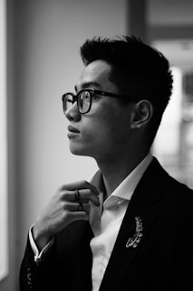 A black and white profile portrait of a person wearing glasses and a suit, with a thoughtful expression. The focus is on the subject's face and hand, adjusting their collar.