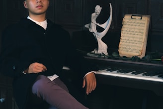 A person dressed in a dark coat sits next to a piano. On the piano, there is a sculpture and a sheet of music. The setting appears to be dimly lit, creating a calm and introspective atmosphere.