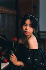 A woman in a black off-the-shoulder dress with sequined details holds a red rose while standing beside a piano. The setting appears to be a dimly lit room with wooden walls and a decorative frame. Her expression is calm and contemplative.