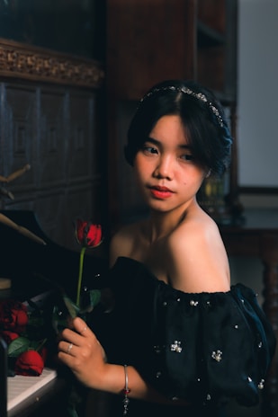 A woman in a black off-the-shoulder dress with sequined details holds a red rose while standing beside a piano. The setting appears to be a dimly lit room with wooden walls and a decorative frame. Her expression is calm and contemplative.