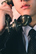A close-up view of a person holding an old-fashioned rotary phone headset near their mouth, dressed in a formal suit and polka-dotted tie. The image captures the lower half of the face and emphasizes the vintage style of the telephone.