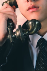A close-up view of a person holding an old-fashioned rotary phone headset near their mouth, dressed in a formal suit and polka-dotted tie. The image captures the lower half of the face and emphasizes the vintage style of the telephone.