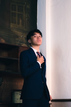 A young person dressed in a formal blue suit and red striped tie stands confidently against a plain wall. The setting includes an old-fashioned rotary phone and a file cabinet, suggesting a vintage office atmosphere.
