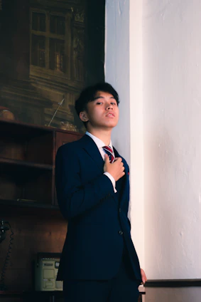 A young person dressed in a formal blue suit and red striped tie stands confidently against a plain wall. The setting includes an old-fashioned rotary phone and a file cabinet, suggesting a vintage office atmosphere.