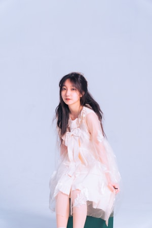 A young woman is seated and wearing a light, sheer dress with delicate detailing. Her long dark hair contrasts with the soft pastels of her outfit, which blends into the light background.