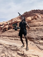 A person stands on rocky terrain, appearing enthusiastic or shouting, while holding a professional camera rig with a microphone. The background consists of layered rock formations under a partly cloudy sky.
