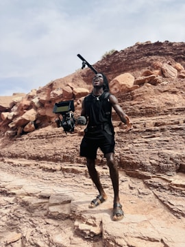A person stands on rocky terrain, appearing enthusiastic or shouting, while holding a professional camera rig with a microphone. The background consists of layered rock formations under a partly cloudy sky.