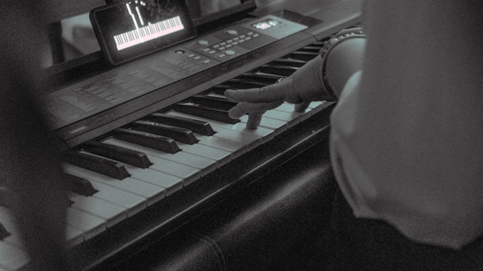 A person plays a keyboard, with one hand positioned over the keys. A tablet is placed above the keyboard displaying sheet music or a virtual keyboard interface.