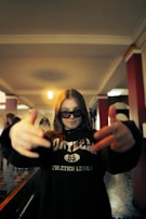 A person with long hair is wearing dark sunglasses and a sweatshirt with the word 'athletics' on it. They have their hands stretched outwards towards the camera, creating a dynamic and energetic pose. The background features an indoor setting with red and white pillars and a patterned floor.
