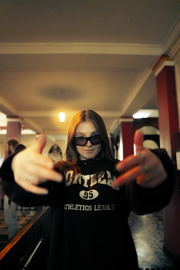 A person with long hair is wearing dark sunglasses and a sweatshirt with the word 'athletics' on it. They have their hands stretched outwards towards the camera, creating a dynamic and energetic pose. The background features an indoor setting with red and white pillars and a patterned floor.