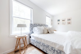 A bright and airy bedroom featuring a bed with a patterned blue and white headboard and fluffy white bedding. On one side, there is a small wooden side table with a sleek, modern lamp. The room has two large windows allowing natural light to flood in, and framed pictures are neatly arranged on one wall. The wooden floor adds a warm tone to the serene atmosphere.