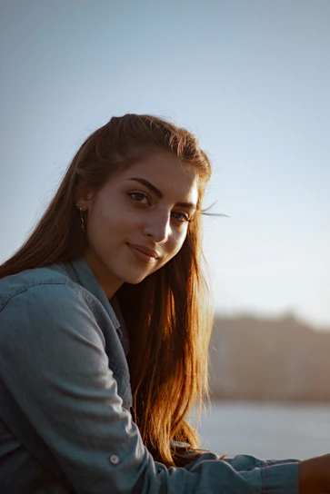 A stylish woman wearing a light blue denim shirt, standing outdoors with natural sunlight.