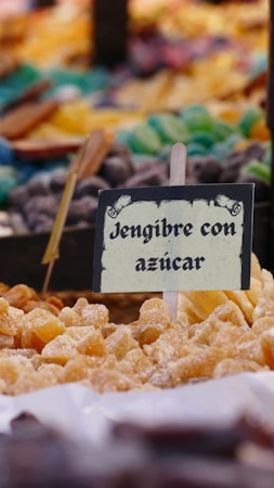 A market or stall display featuring a variety of colorful candies and sweets, with a sign in the foreground labeled 'Jengibre con az&uacute;car' indicating ginger with sugar. The focus is primarily on the sugared ginger pieces that are at the front of the display, with other confections and candies visible but blurred in the background.
