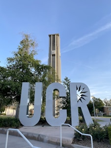 A large metallic sculpture of the letters 'UCR' is positioned in the foreground, surrounded by greenery and pathways. Behind it stands a tall clock tower, with trees and a clear blue sky in the background. The architecture is modern and well-maintained.