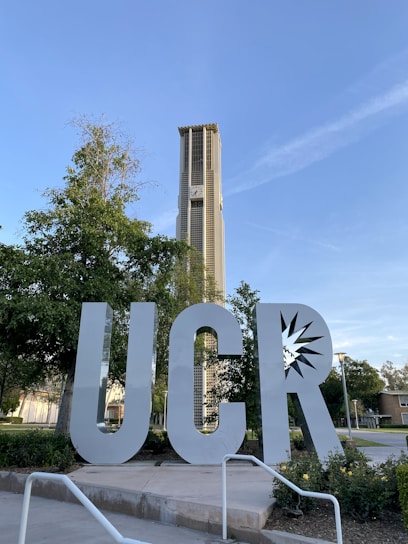 A large metallic sculpture of the letters 'UCR' is positioned in the foreground, surrounded by greenery and pathways. Behind it stands a tall clock tower, with trees and a clear blue sky in the background. The architecture is modern and well-maintained.