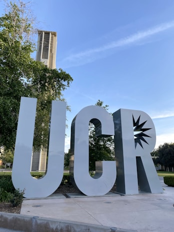 Large, metallic letters 'UCR' are prominently displayed in an outdoor setting. Behind the letters, there is a tall tower structure visible through the foliage of the surrounding trees. The sky is clear with a few wispy clouds, suggesting fair weather.