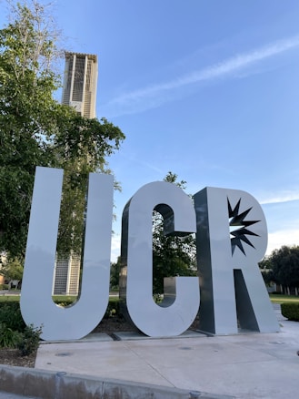 Large, metallic letters 'UCR' are prominently displayed in an outdoor setting. Behind the letters, there is a tall tower structure visible through the foliage of the surrounding trees. The sky is clear with a few wispy clouds, suggesting fair weather.