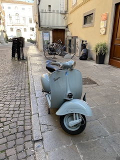 A pale blue vintage Vespa scooter is parked on a cobblestone street near a building with potted plants and bicycles. There are several chairs stacked near the corner of the building, and the street extends into the background with more bicycles and buildings visible. The scene appears to be in an urban European setting.