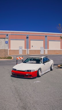 A modified white sports car with a red front bumper is parked on a street in front of a building with a blue sky in the background. The car has lowered suspension and custom wheels.