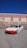 A modified white sports car with a red front bumper is parked on a street in front of a building with a blue sky in the background. The car has lowered suspension and custom wheels.