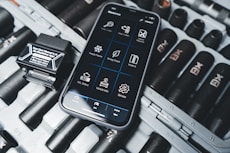 A smartphone displaying an application interface with various diagnostic tools is placed on a surface scattered with dark cylindrical objects resembling large batteries or tools. Beside the phone is a small black device with a label that includes text and a phone number.