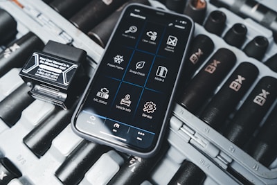 A smartphone displaying an application interface with various diagnostic tools is placed on a surface scattered with dark cylindrical objects resembling large batteries or tools. Beside the phone is a small black device with a label that includes text and a phone number.