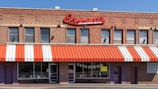 A brick building houses a barbecue restaurant with a red and white striped awning. Large red signage displays the name 'Bryant's Barbeque' above the windows. There are multiple windows with a few signs and posters visible. The main entrance is flanked by additional doors and brick walls.
