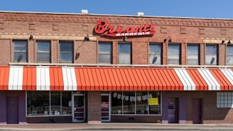 A brick building houses a barbecue restaurant with a red and white striped awning. Large red signage displays the name 'Bryant's Barbeque' above the windows. There are multiple windows with a few signs and posters visible. The main entrance is flanked by additional doors and brick walls.