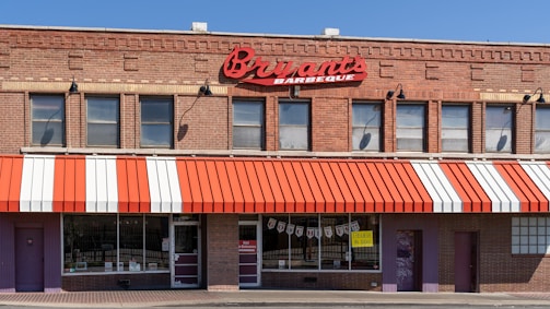 A brick building houses a barbecue restaurant with a red and white striped awning. Large red signage displays the name 'Bryant's Barbeque' above the windows. There are multiple windows with a few signs and posters visible. The main entrance is flanked by additional doors and brick walls.