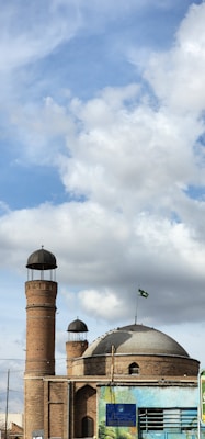 A historic brick building features a large dome and two tall, cylindrical minarets, topped with black domes. A green flag with white symbols is mounted on a pole extending from the dome. Fluffy white clouds and a bright blue sky provide a picturesque backdrop. The lower section of the image shows an ornate mural painted on the structure, adding vibrant colors to the scene.