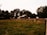 A rustic barn surrounded by a wooden fence sits in the middle of a lush green field. Tall trees create a natural background barrier, filtering the light and casting shadowy areas. The atmosphere suggests a rural and serene environment.