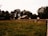 A rustic barn surrounded by a wooden fence sits in the middle of a lush green field. Tall trees create a natural background barrier, filtering the light and casting shadowy areas. The atmosphere suggests a rural and serene environment.
