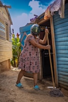 A person wearing a helmet and a patterned dress is seen mopping the ground outside a wooden house. The house is blue and brown with laundry hanging near the entrance. The person is wearing blue slippers and there are plants visible in the background under a clear blue sky.