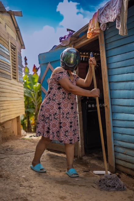 A person wearing a helmet and a patterned dress is seen mopping the ground outside a wooden house. The house is blue and brown with laundry hanging near the entrance. The person is wearing blue slippers and there are plants visible in the background under a clear blue sky.