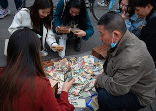 A group of people are gathered around a collection of vintage postcards or trading cards laid out on the ground. They appear to be inspecting the items with interest. Some individuals are crouched down, while others are kneeling, and there is a bicycle wheel visible in the background.