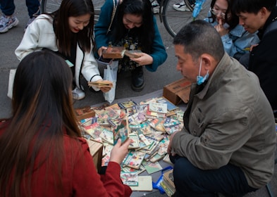 A group of people are gathered around a collection of vintage postcards or trading cards laid out on the ground. They appear to be inspecting the items with interest. Some individuals are crouched down, while others are kneeling, and there is a bicycle wheel visible in the background.