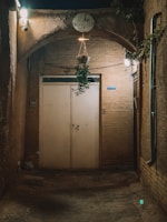 An entryway to a small, rustic cafe with a hanging plant above the door. The area is dimly lit by two lights on either side, creating a cozy atmosphere. The walls are made of brick and stone, giving it an old-world charm. A sign above the door reads 'Simaar Cafe'.