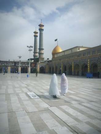 A large mosque courtyard with two people wearing traditional garments walking towards a structure featuring ornate architectural elements, including a golden dome and two tall minarets. The sky is overcast with scattered clouds.