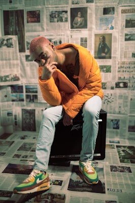 A person with dyed hair is sitting on a black amplifier. They are wearing a bright orange puffer jacket and light blue jeans. The sneakers are colorful with green, brown, yellow, and white elements. The setting is covered with newspaper pages on the walls and floor, creating a distinctive backdrop.