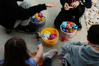 Several children are sitting on the ground with buckets filled with colorful plastic eggs and toys. The surrounding area is covered in concrete and rocks. One child is extending their arm with an open hand, possibly in conversation or to show the items collected. The image portrays a casual and playful setting.