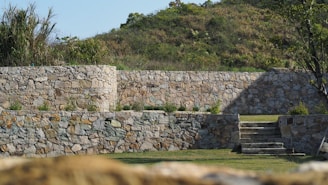 Stone walls create a terraced effect in a landscaped garden, with stairs leading up to the higher levels. The walls are constructed from irregularly shaped stones with various shades of gray and tan. In the background, there is a small hill covered with greenery and bushes, and a tree is visible on the right side.