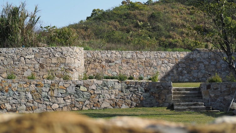 Stone walls create a terraced effect in a landscaped garden, with stairs leading up to the higher levels. The walls are constructed from irregularly shaped stones with various shades of gray and tan. In the background, there is a small hill covered with greenery and bushes, and a tree is visible on the right side.