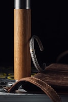 A wooden cylindrical object with a metallic top stands upright. A detailed silver bracelet with a textured design hangs on the wood. In the foreground, a brown leather strap with stitching is partially visible, alongside what appears to be the edge of a watch case. A leather-bound book or notebook is positioned in the background. The setting is dimly lit, creating a rich contrast among the textures and materials.